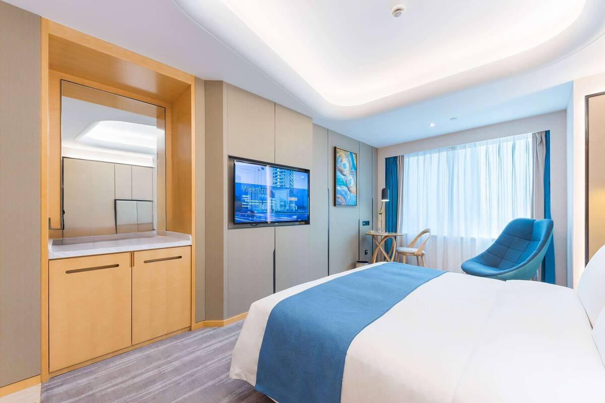 A modern hotel room with a large bed, a television mounted on the wall, a small table with a chair, and a mirror in the corner.
