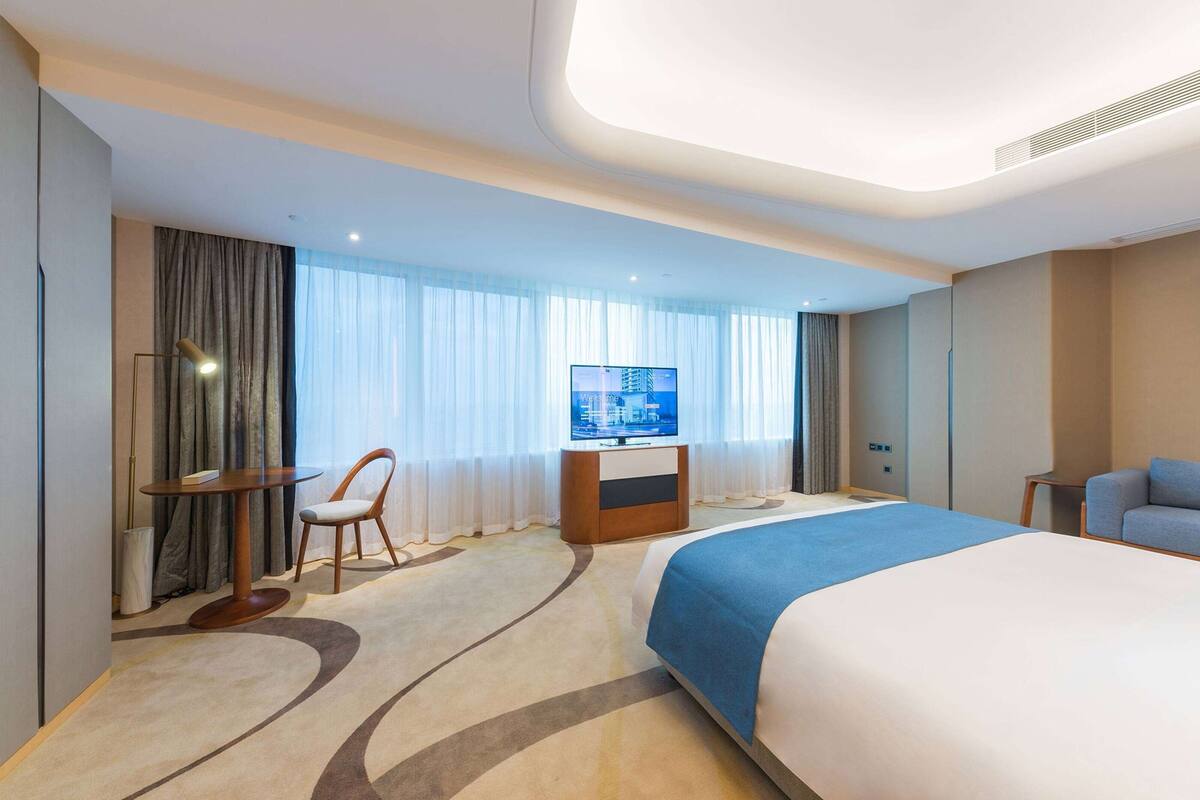 A modern hotel room with a large bed, a television, a desk with a chair, and a sofa.