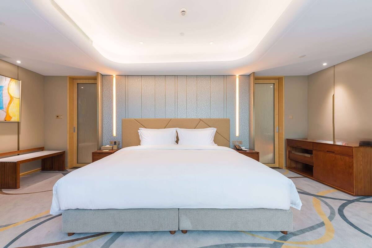 A modern hotel room with a large bed, bedside tables, a wooden bench, a desk, and a wardrobe.