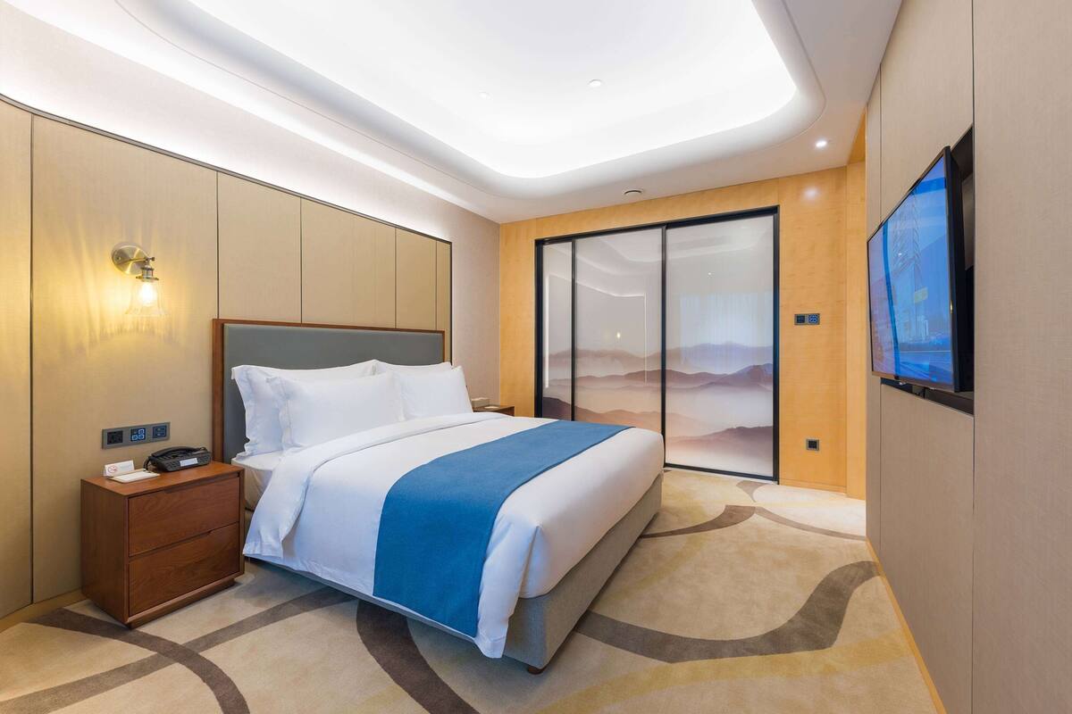 A modern hotel room with a large bed, a nightstand, a flat-screen TV, and a sliding glass door leading to a balcony with a desert mural.