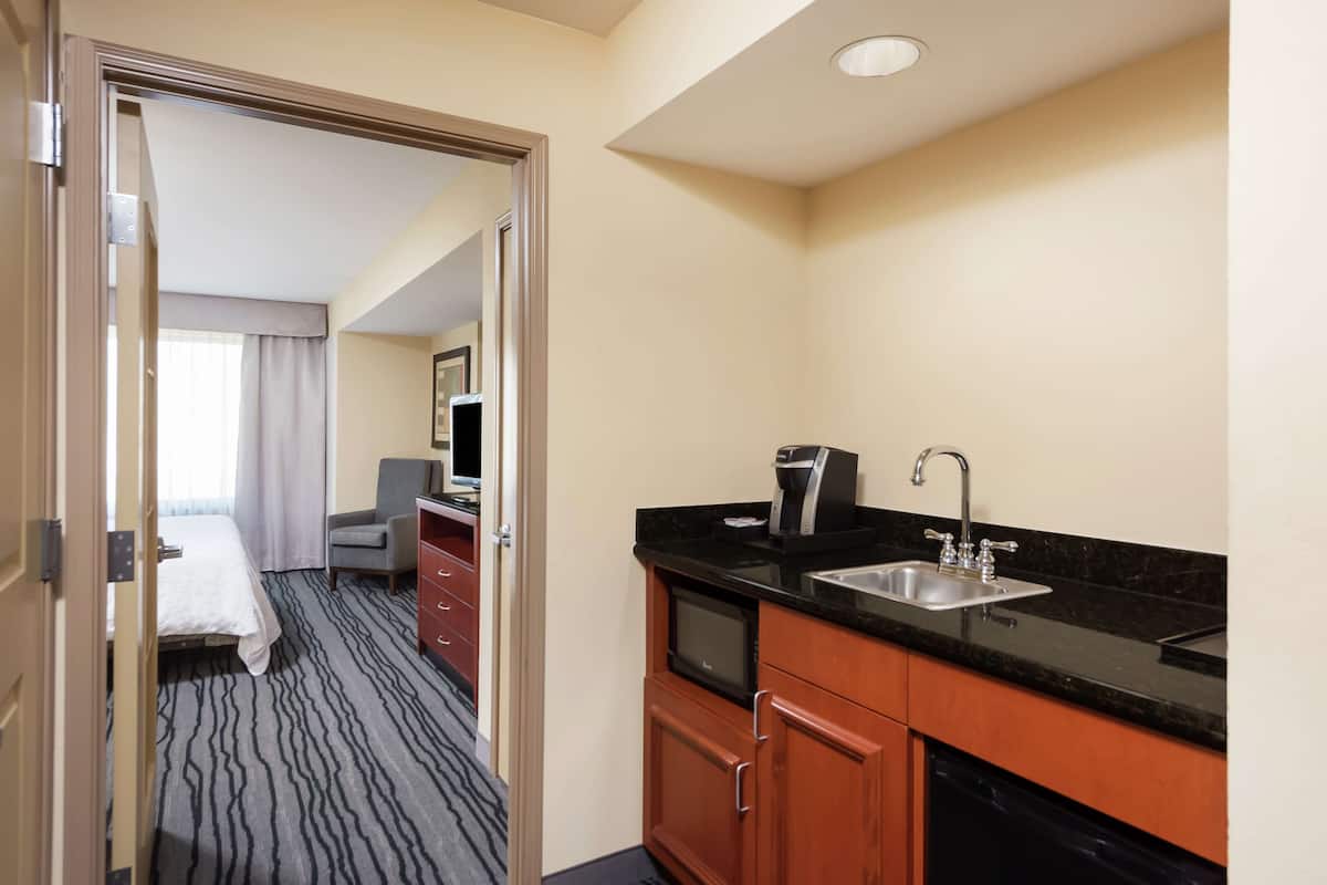 A hotel room with a bed, a desk, a chair, a kitchenette with a sink and coffee maker, and a bathroom with a toilet and sink.