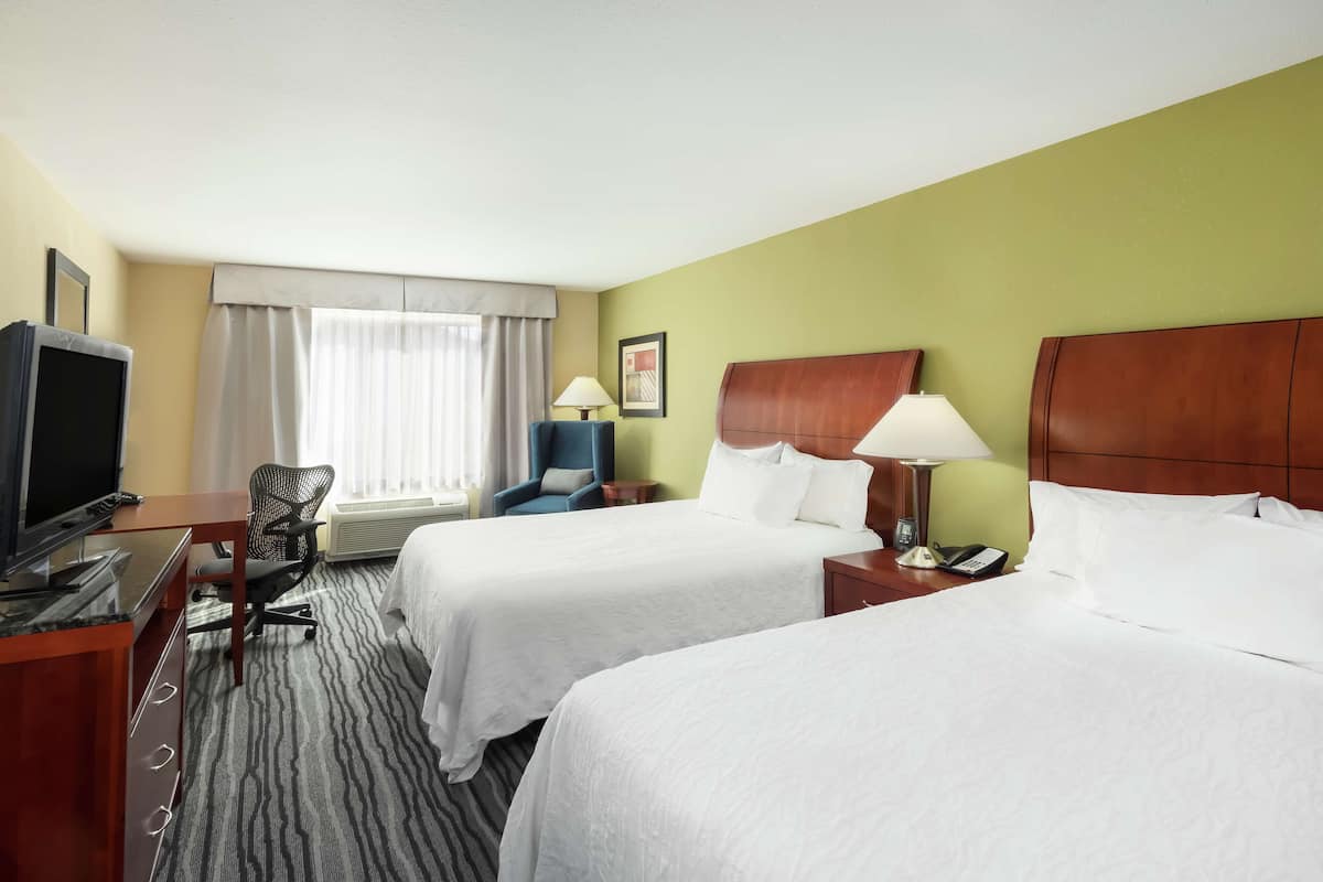 A hotel room with two beds, a desk with a television, a chair, and a window with curtains.