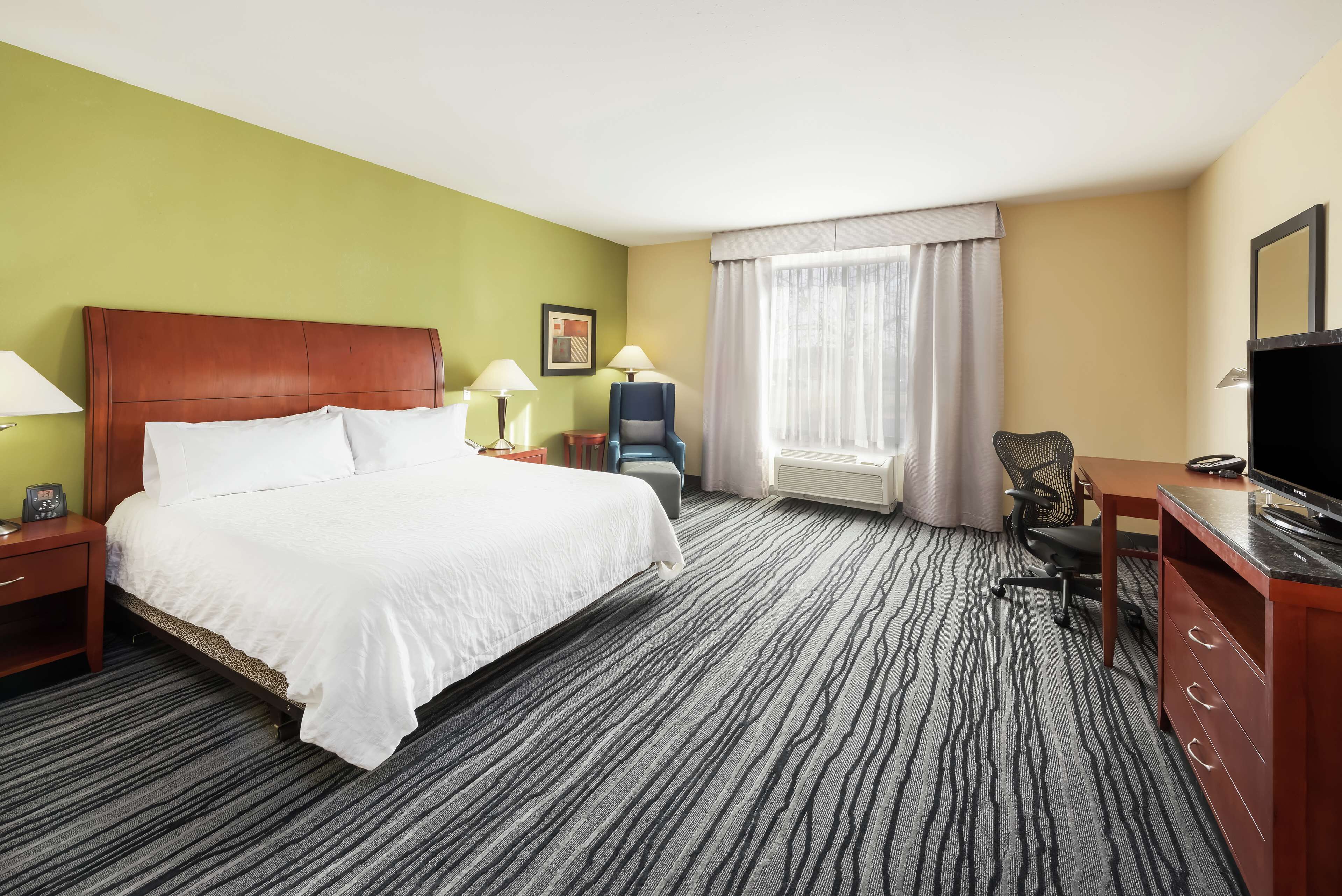 A hotel room with a large bed, two bedside tables with lamps, a desk with a chair, a television, and a window with curtains.