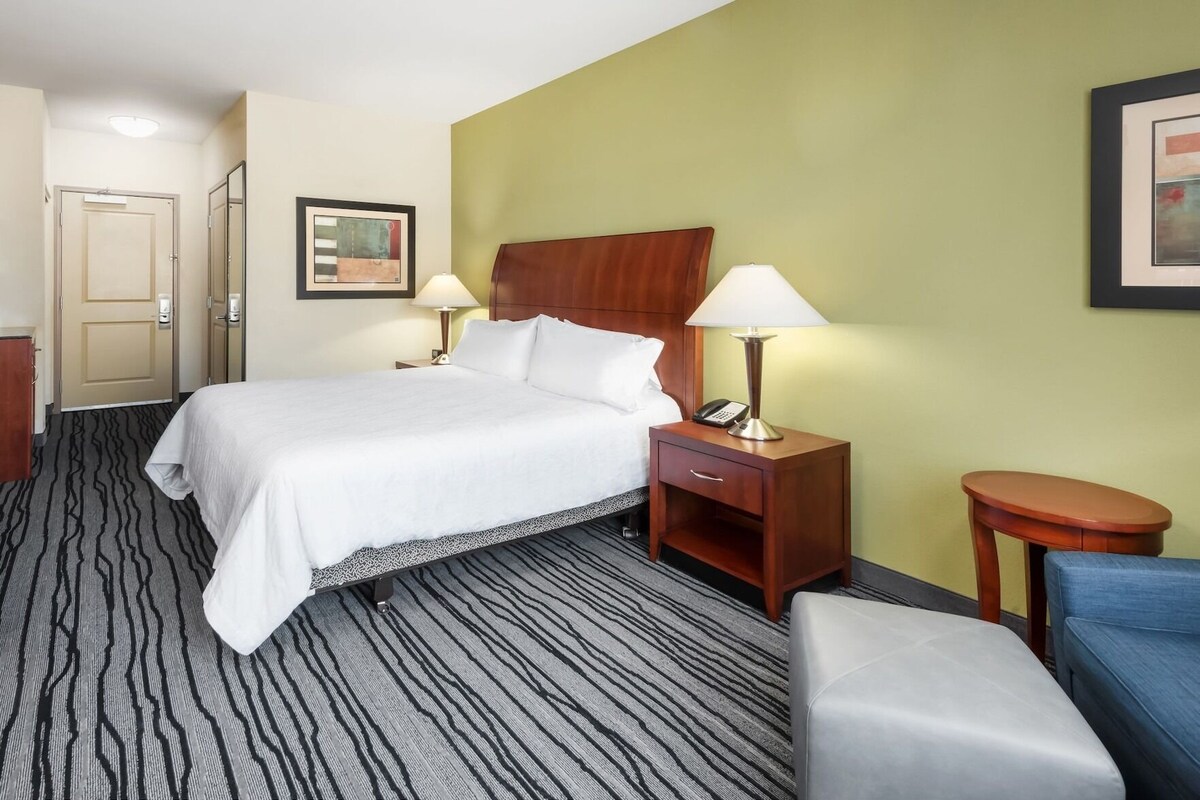 Una habitación de hotel con una cama, dos mesitas de noche con lámparas, una silla y una puerta.