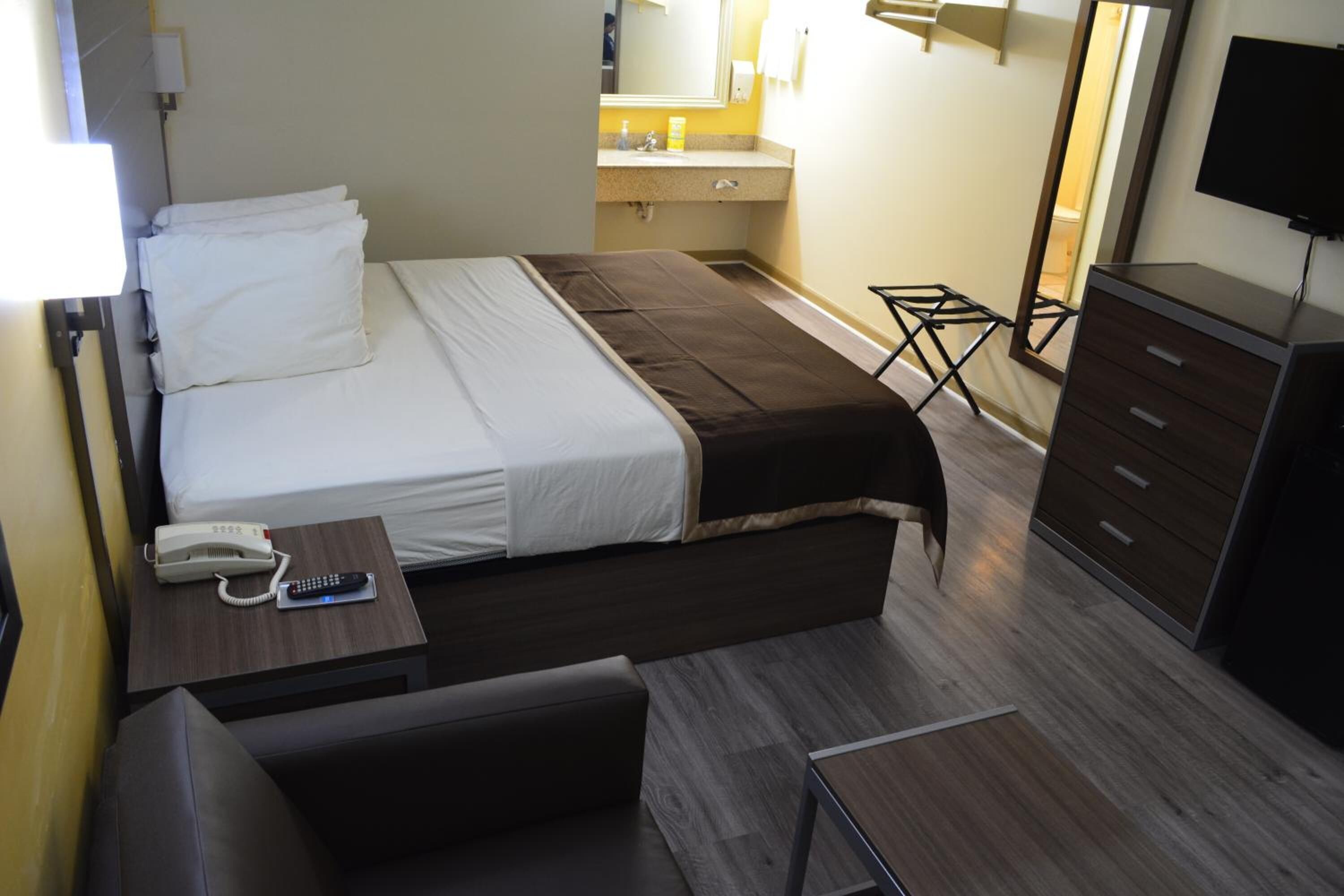 Quarto de hotel com cama, mesa de cabeceira com telefone e controle remoto, um pequeno espaço para sentar, cômoda e espelho.