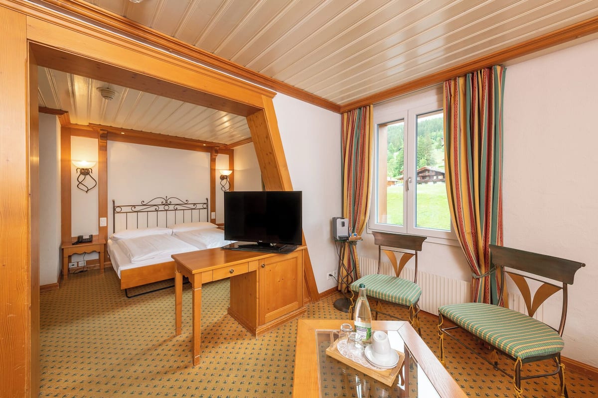 Ein Zimmer mit zwei Betten, einem Fernseher, einem Tisch mit einer Flasche und Gläsern sowie einem Fenster mit Vorhängen.