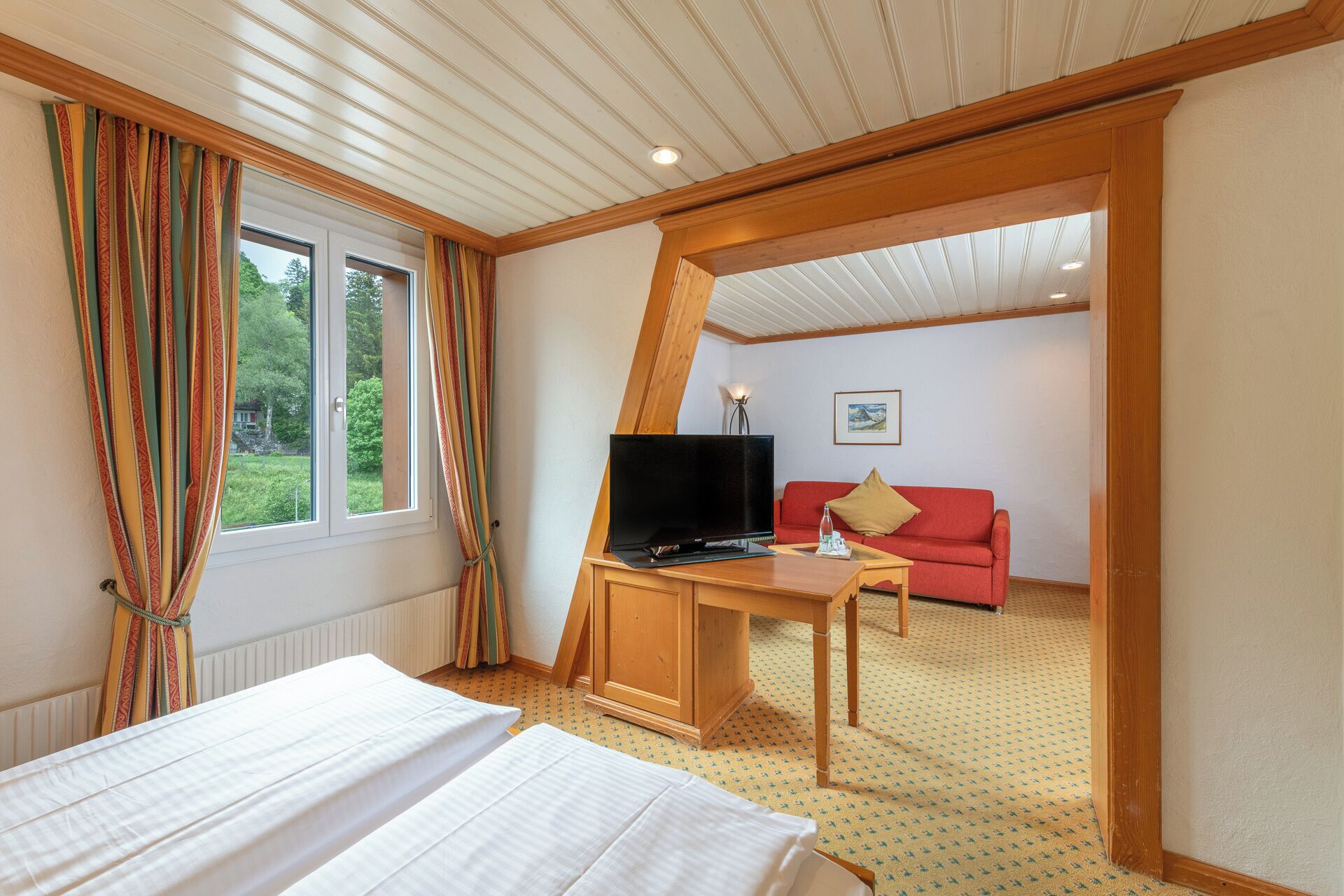 Una habitación de hotel con cama, escritorio con televisor, sofá rojo y ventana con cortinas.