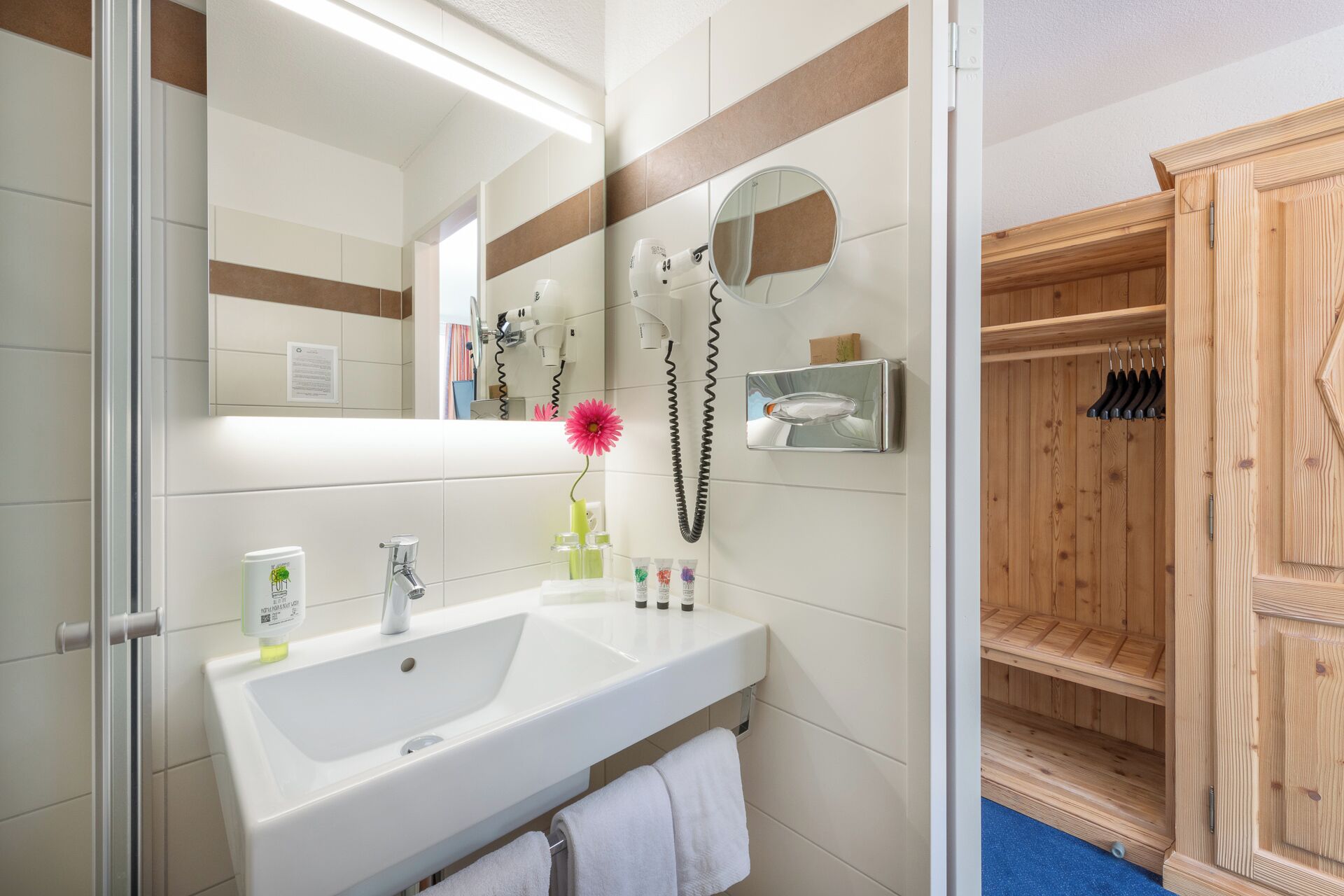 Baño con lavamanos blanco, espejo y toallero. Se ve una sauna de madera a través de una puerta entreabierta.
