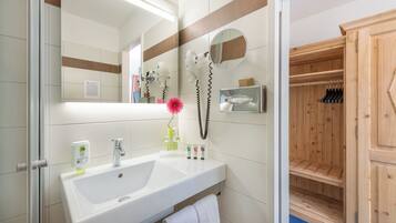 Baño con lavamanos blanco, espejo y toallero. Se ve una sauna de madera a través de una puerta entreabierta.
