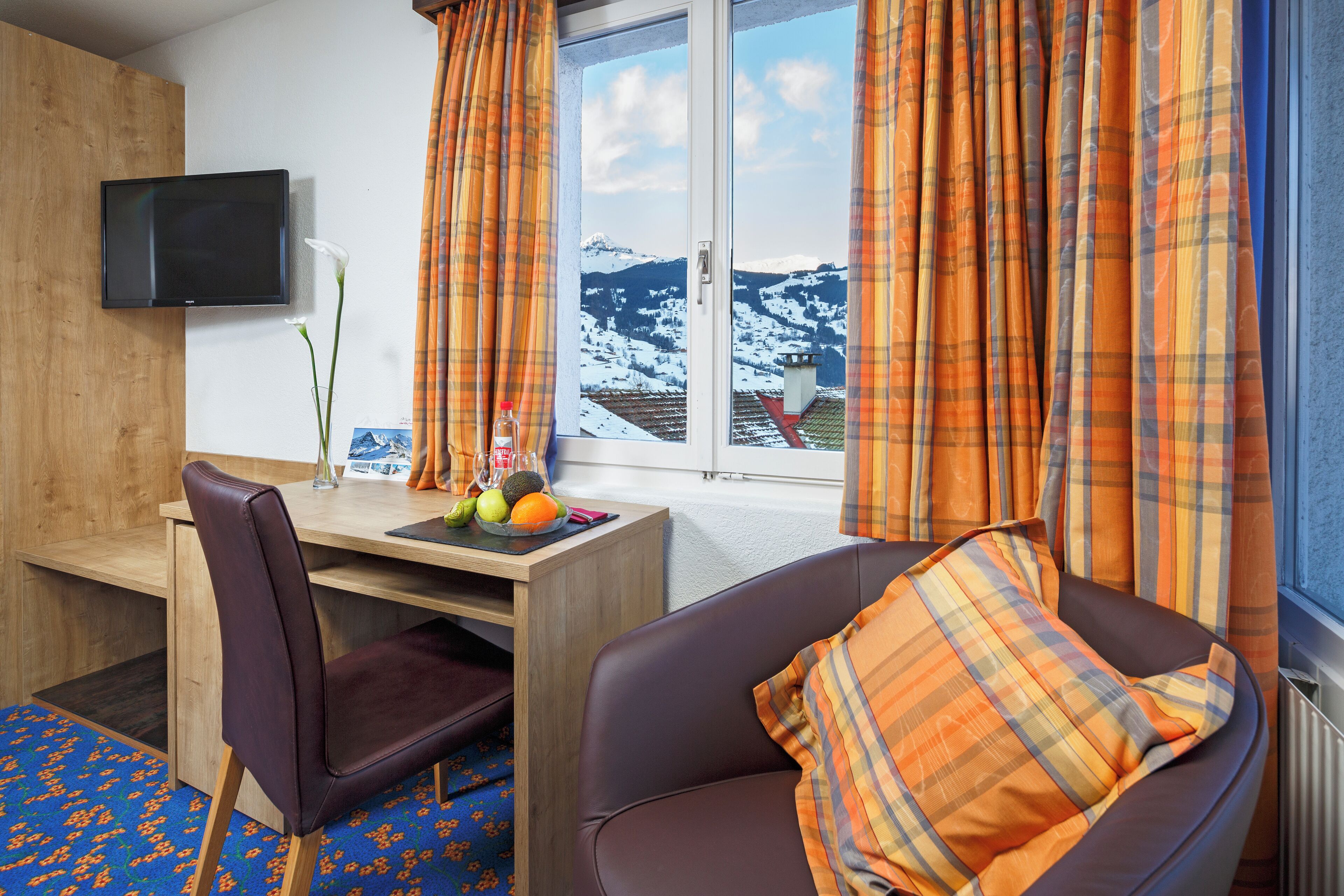 Ein Hotelzimmer mit Schreibtisch aus Holz, einem an der Wand befestigten Fernseher, einem bequemen Sessel mit Karokissen und einem Fenster mit Vorhängen, das einen Blick auf schneebedeckte Berge bietet.