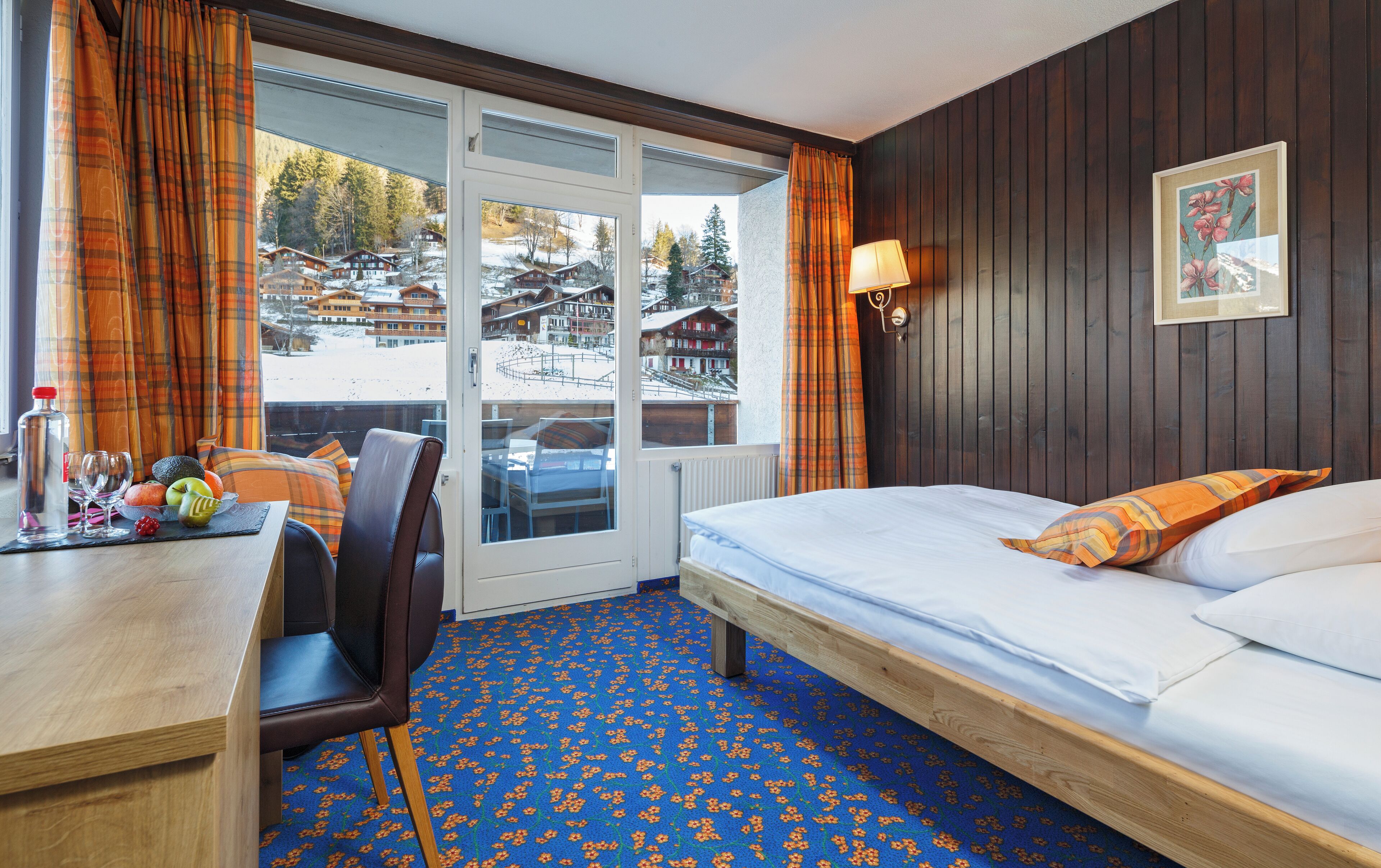 A hotel room with a bed, a desk with a chair, a table with a bottle and glasses, and a view of a snowy landscape.