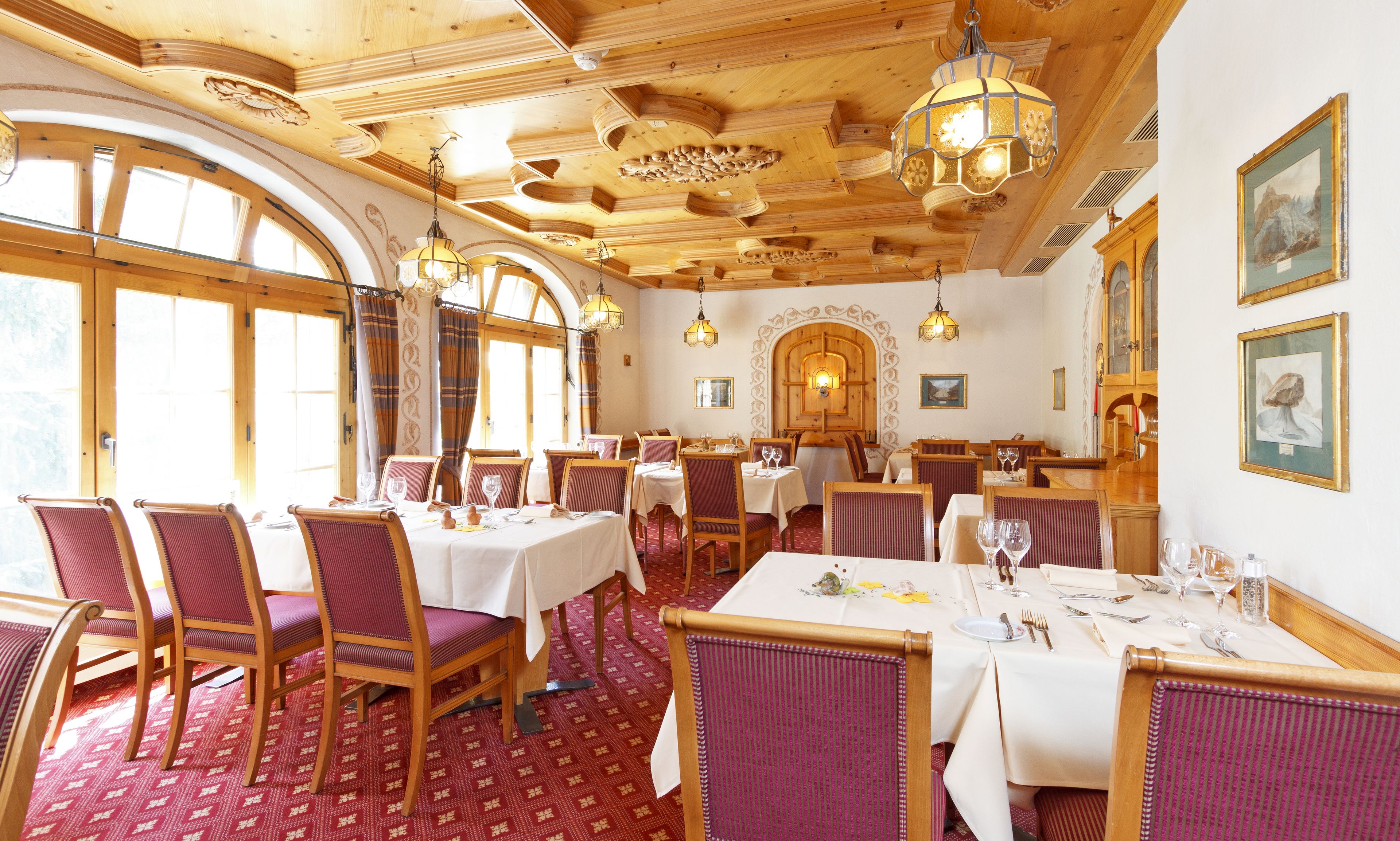 Un restaurante con techos de madera, ventanas amplias y alfombra roja estampada. Las mesas están dispuestas con manteles blancos y sillas rojas.