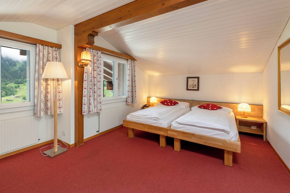 Ein Schlafzimmer mit zwei Betten, einem fensterbelaubten Fenster, einer Lampe und Blick auf eine grüne Landschaft.
