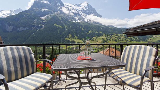 A balcony with two striped chairs and a table, overlooking a mountainous landscape with a snow-capped peak. - Derby Swiss Quality Hotel (Grindelwald)