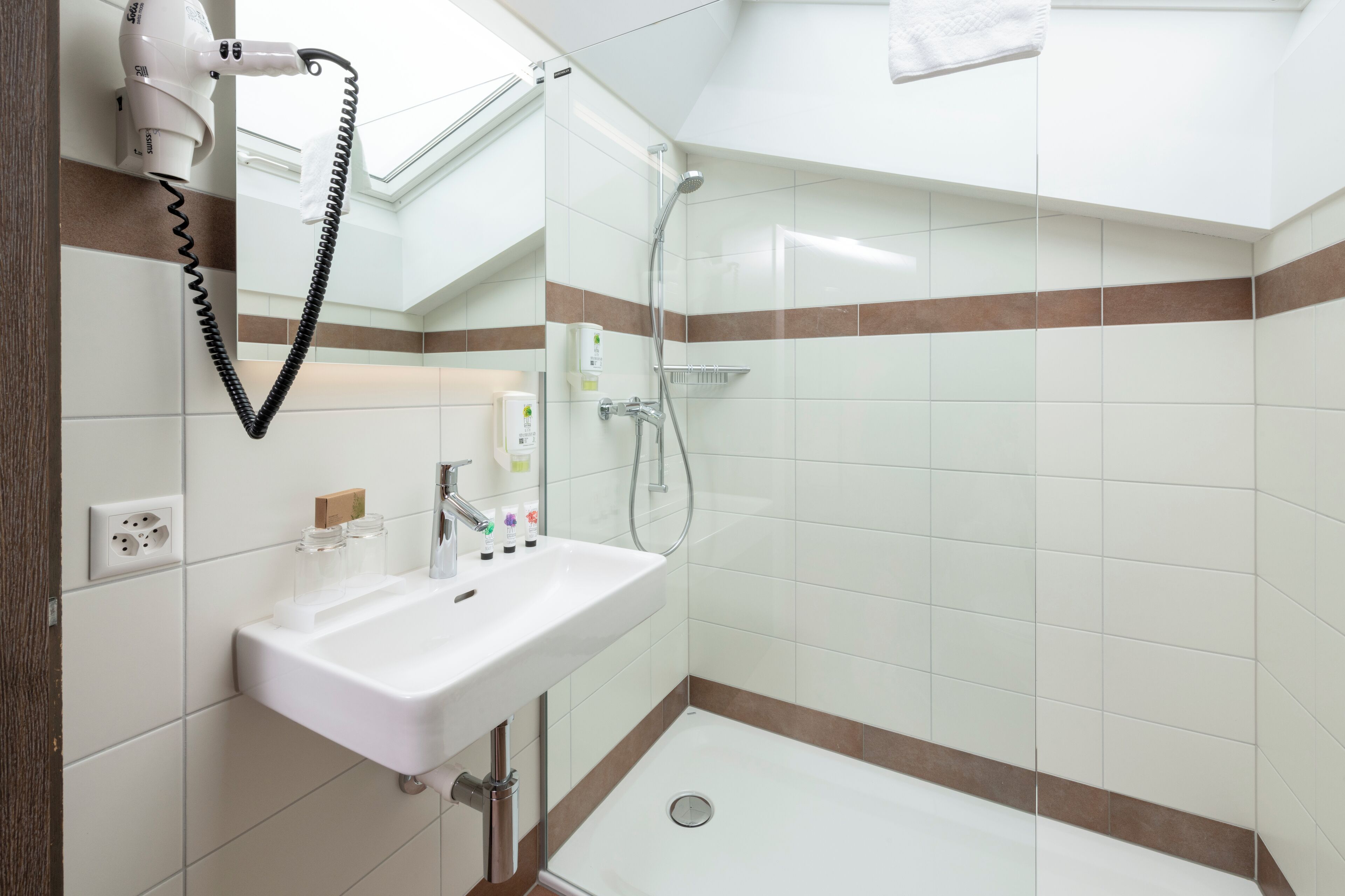 A modern bathroom with a white sink, a handheld hair dryer, a shower, and a towel hanging on the wall.
