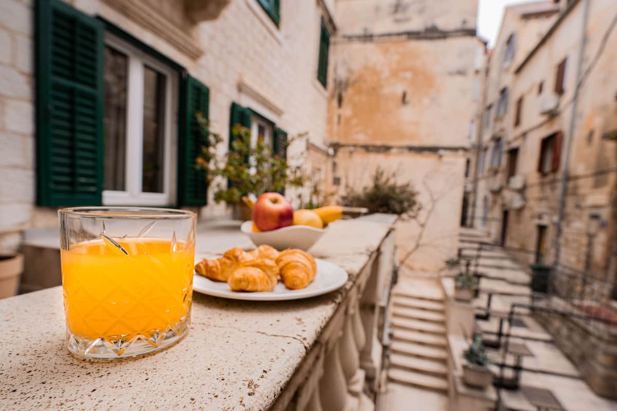 Ein Tisch mit einem Glas Orangensaft, einer Croissant-Platte und einer Obstschale, draußen vor einer Häuserkulisse.