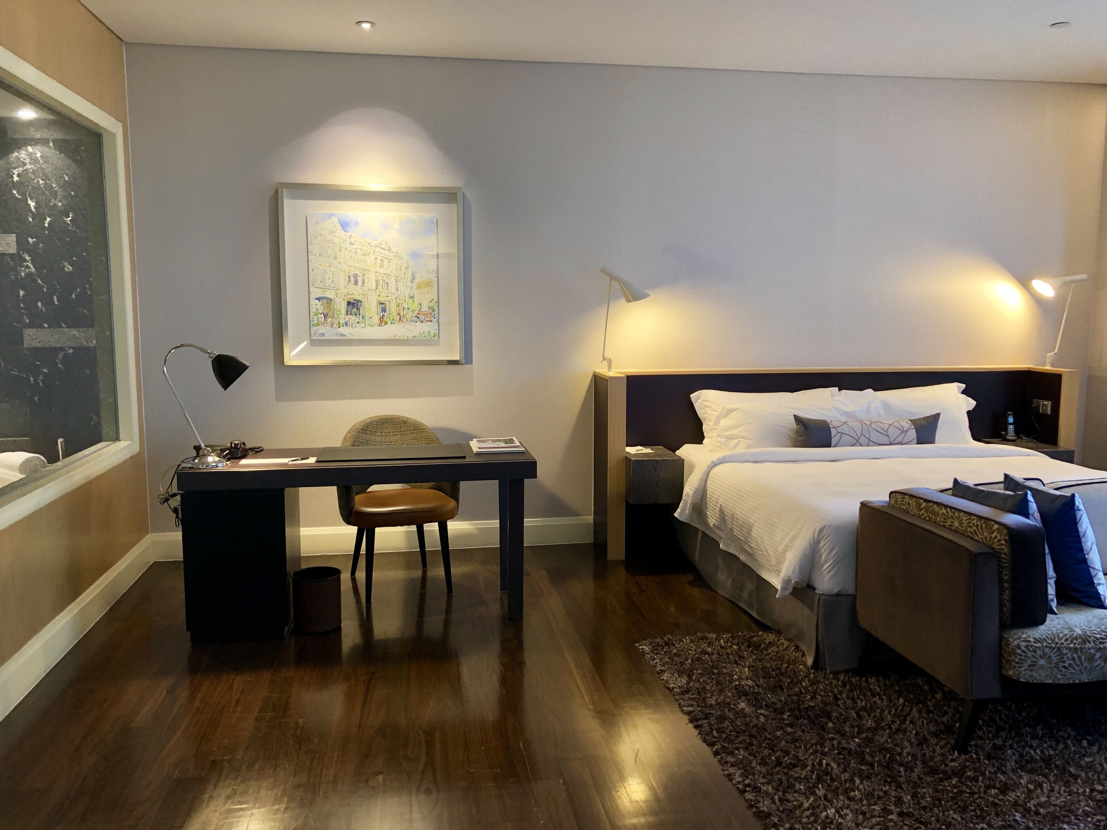 Une chambre d’hôtel moderne avec un plancher en bois, un bureau avec une chaise, un lit avec une literie blanche, une table de chevet, une lampe et un tableau encadré au mur.