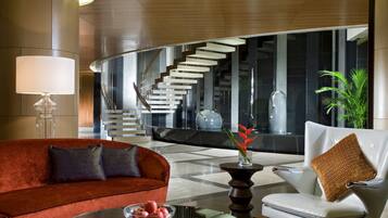 A modern lobby with a red sofa, a white armchair, a glass coffee table with fruit, and a staircase in the background.