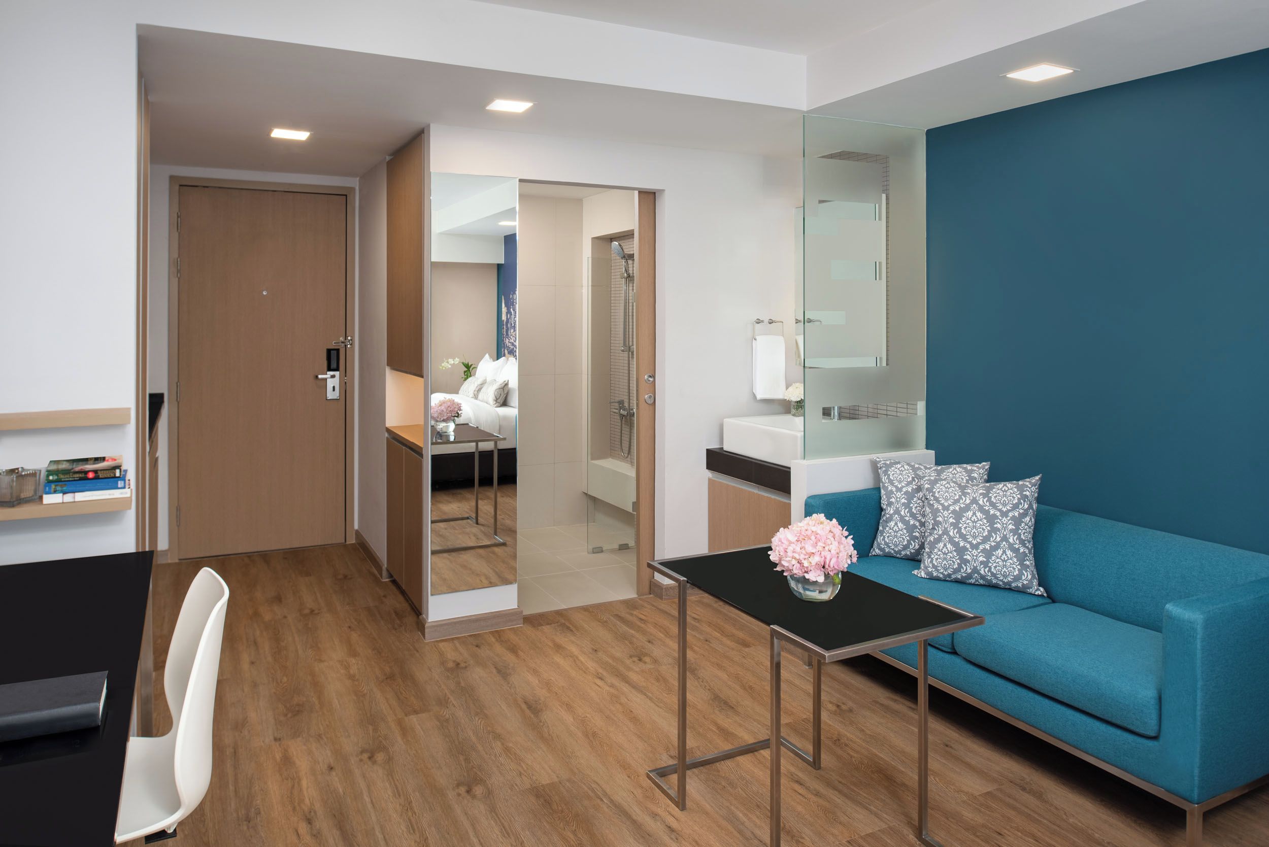 Ein modernes Hotelzimmer mit Holzboden, einem blauen Sofa, einem kleinen Tisch mit einer Vase, einem Spiegel und einer Tür zum Badezimmer.