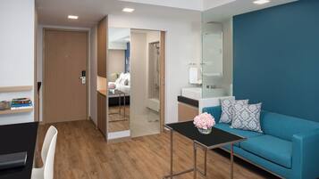 Een moderne hotelkamer met een houten vloer, een blauwe bank, een klein tafeltje met een vaas, een spiegel en een deur naar een badkamer.