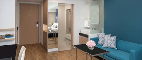 A modern hotel room with a wooden floor, a blue sofa, a small table with a vase, a mirror, and a door leading to a bathroom.