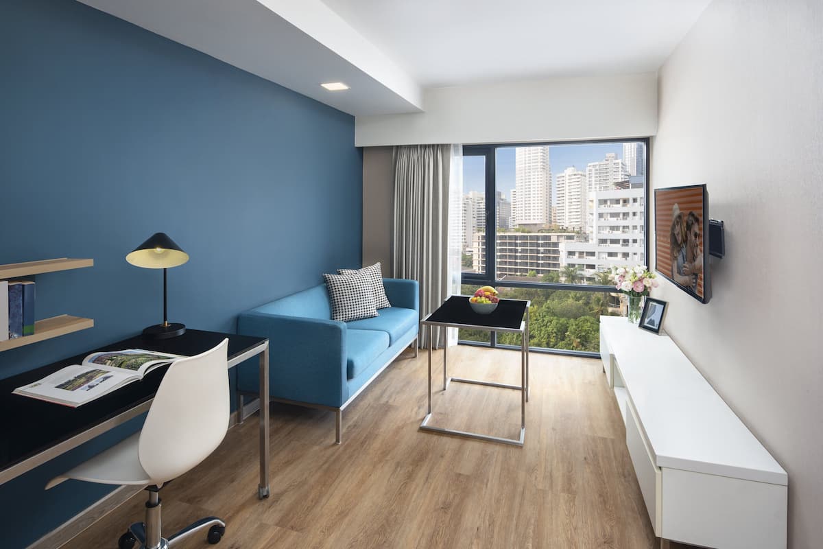 Quarto de hotel moderno com parede azul, sofá, escrivaninha com luminária, mesa pequena com cesta de frutas e uma grande janela com vista para a cidade.