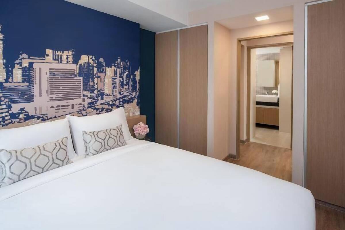 Ein Hotelzimmer mit Bett, einer Stadtlandschaftsmalerei, einem Holzschrank und einem durch eine offene Tür einsehbaren Badezimmer.