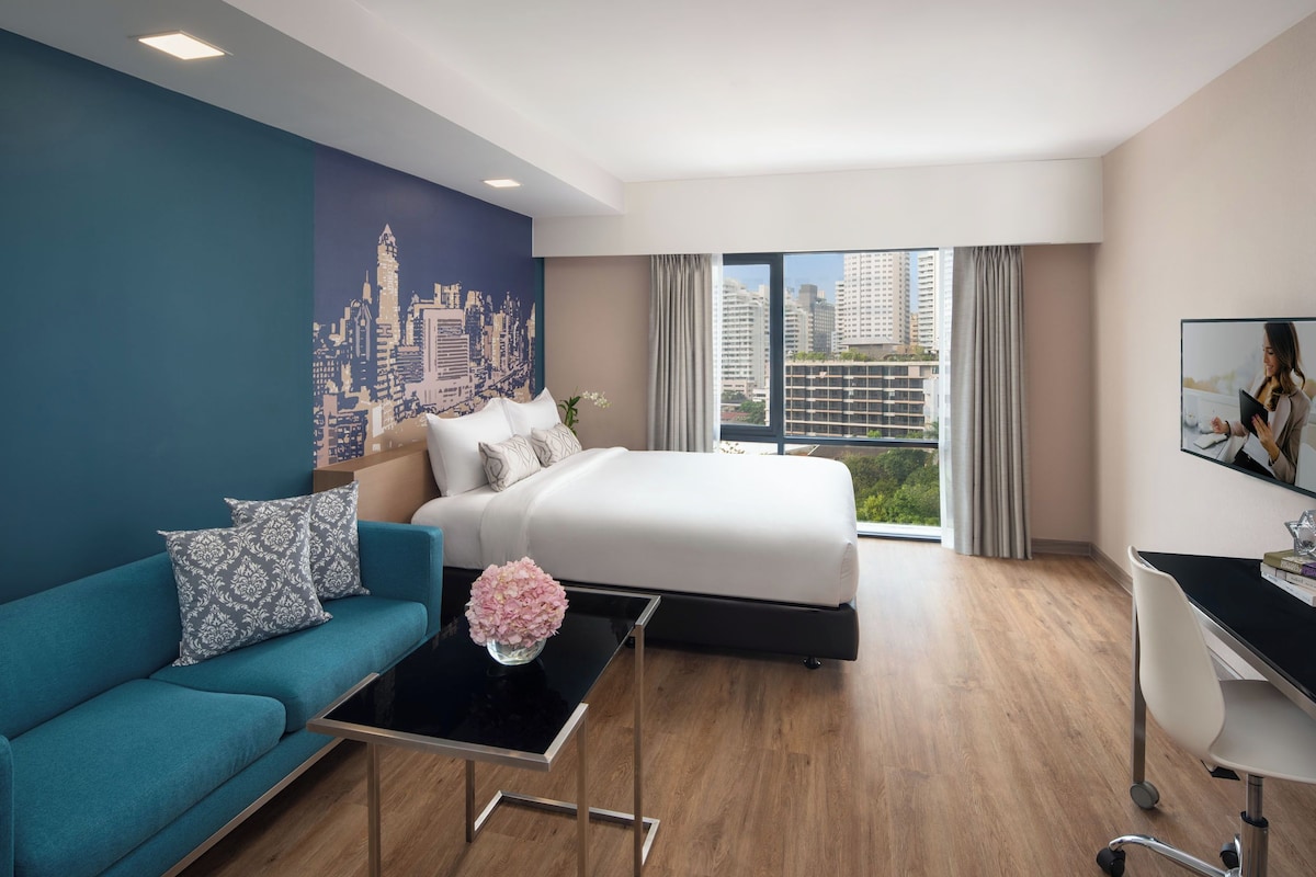 Quarto de hotel moderno com cama, sofá, escrivaninha e vista para a cidade.