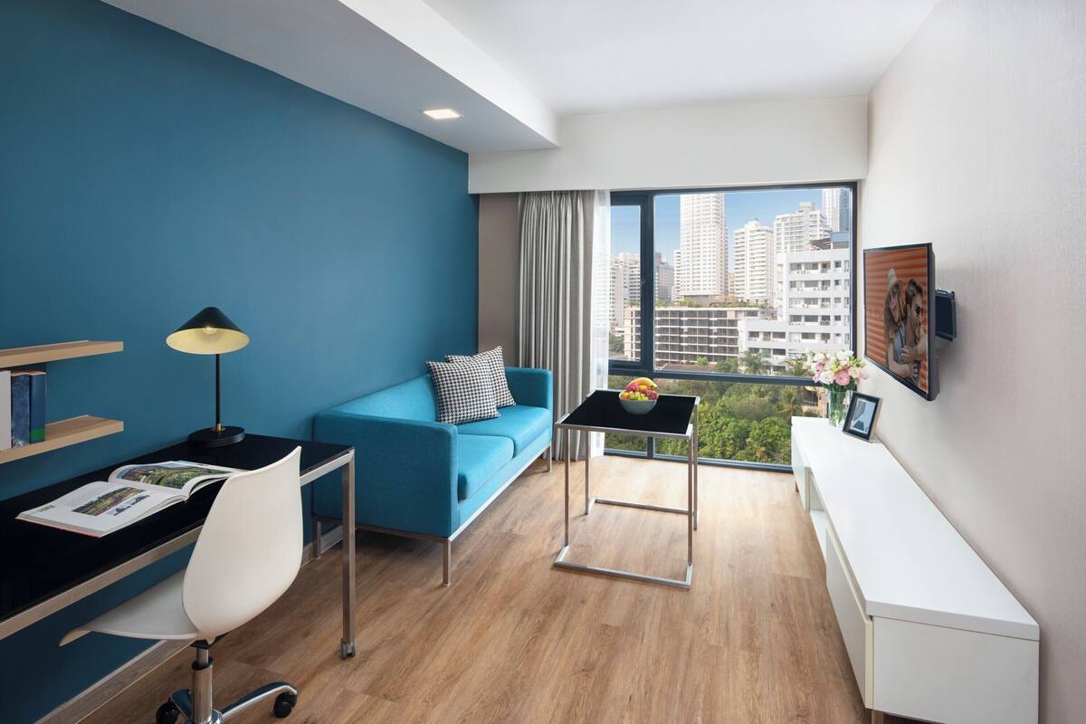Een moderne hotelkamer met een blauwe accentmuur, een bureau met lamp, een sofa, een klein tafeltje met een schaal fruit, en een groot raam met uitzicht op de stad.