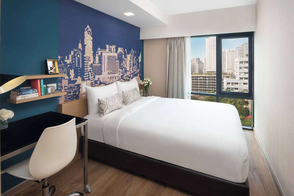 Hotelkamer met stadsmuurschildering, een bed met witte lakens, een bureau met stoel, en een groot raam met uitzicht op gebouwen.