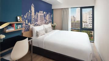 Een hotelkamer met een bed, een bureau, een stoel, een stadslandschap muurschildering en een groot raam met uitzicht op gebouwen.