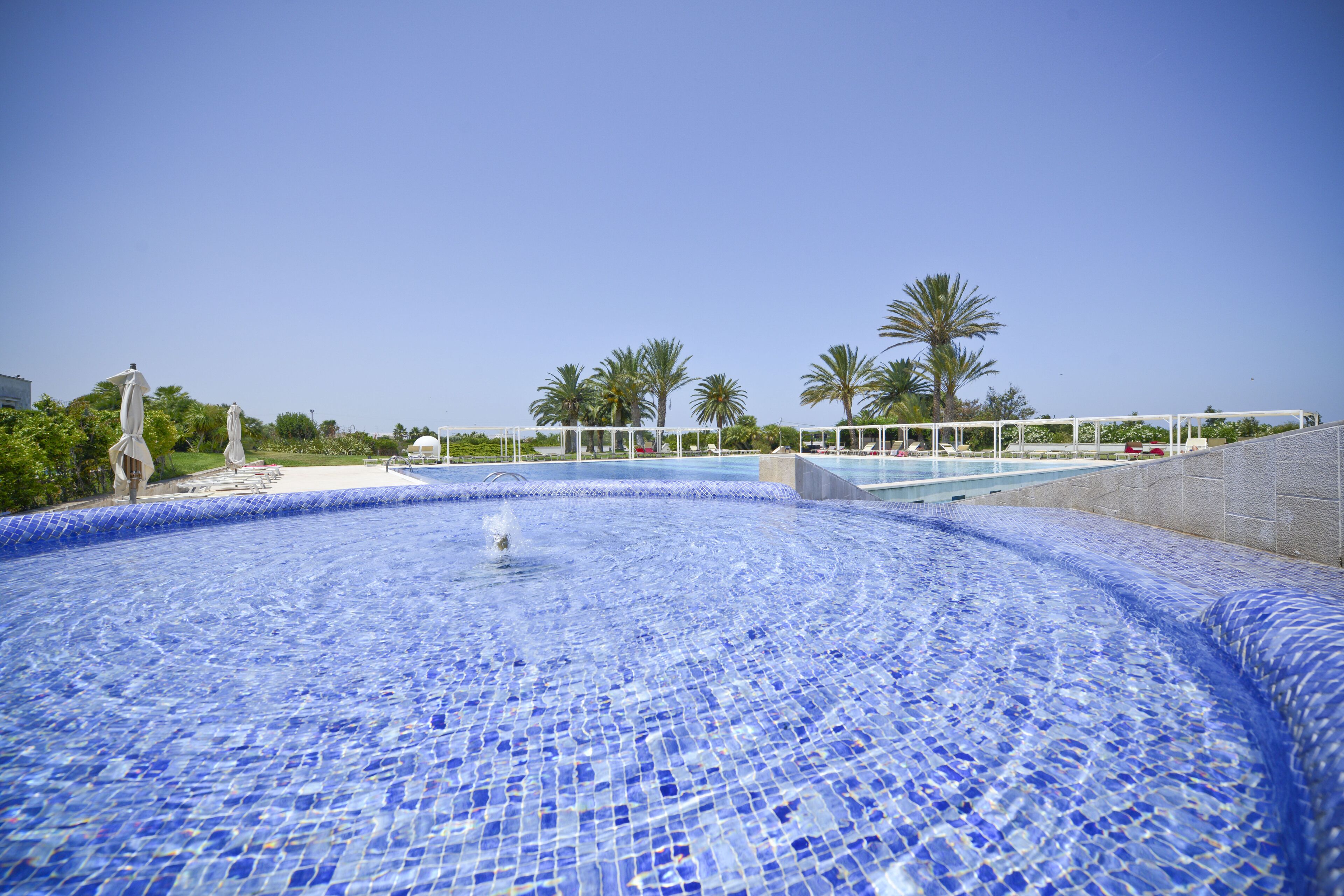 Una piscina con mosaicos, rodeada de palmeras y una valla blanca.