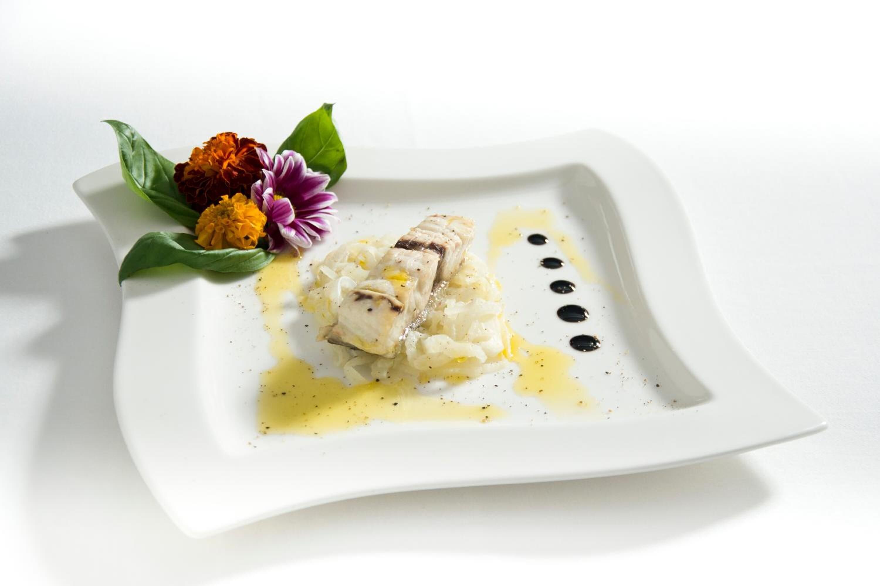 A white square plate with a piece of fish, a flower, and a sauce.