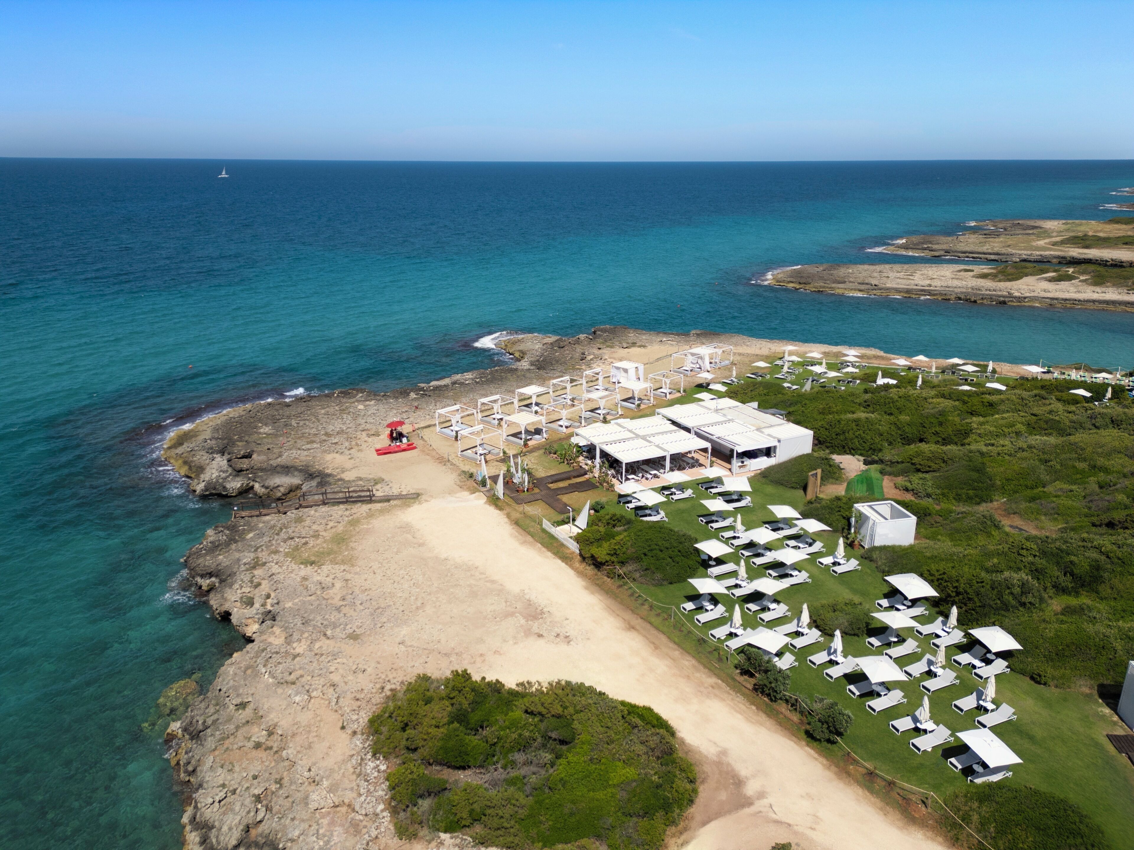 Vista aérea de un complejo turístico frente al mar, con varias construcciones de techo blanco, una playa de arena y una vista despejada del océano.