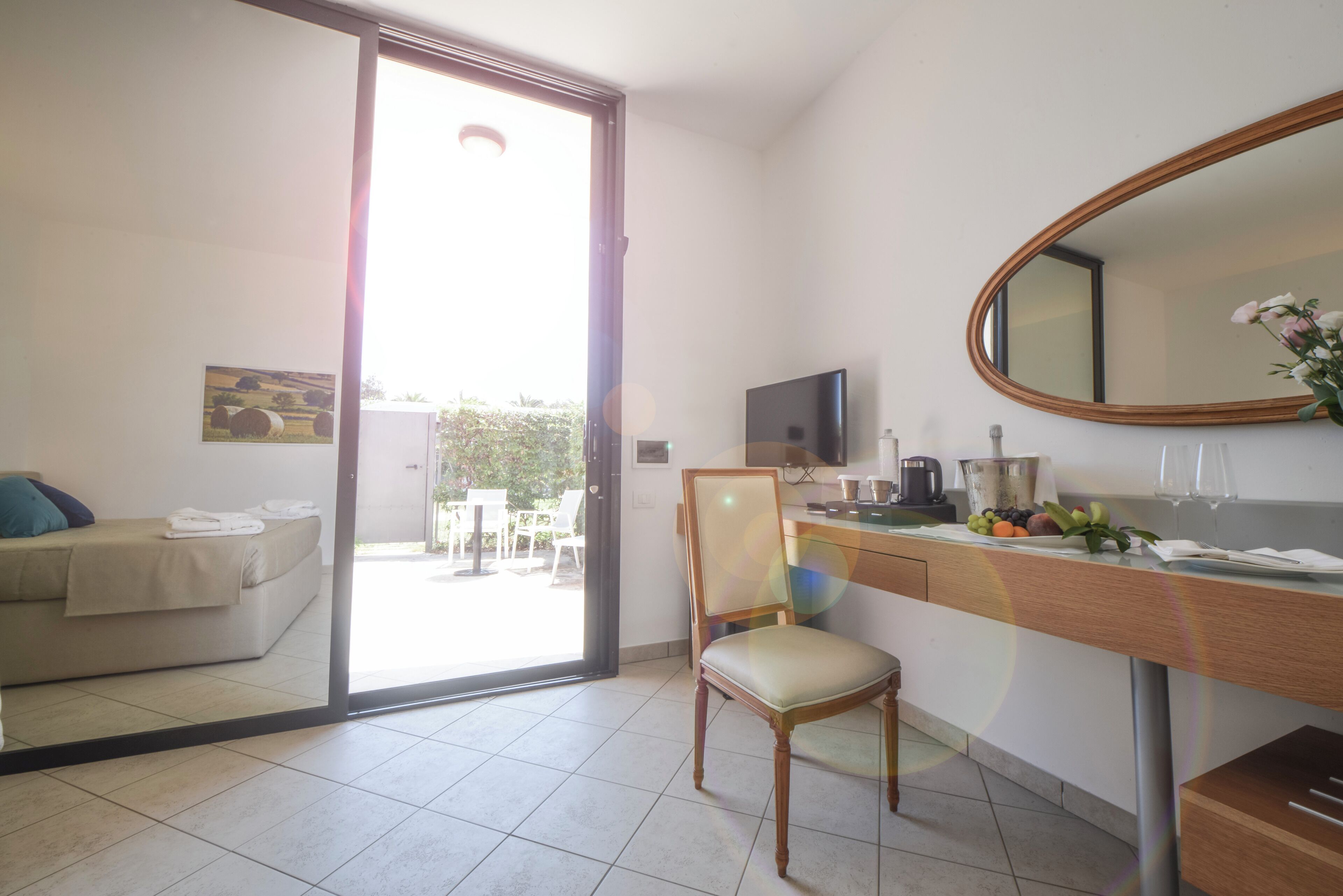 A modern hotel room with a sliding glass door leading to a balcony, a wooden desk with a chair, a television, and a mirror.