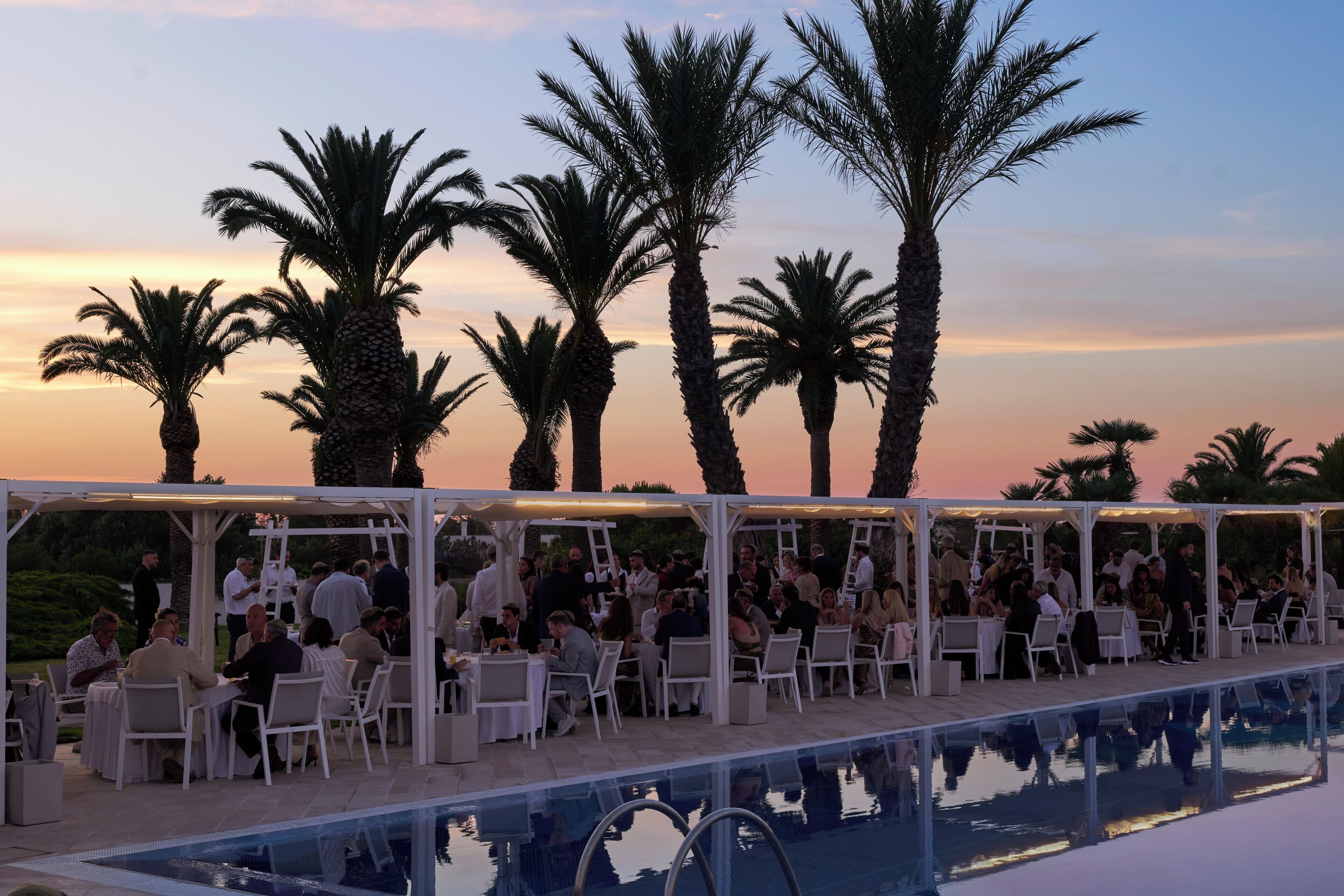 An outdoor pool area with white canopied seating and palm trees in the background.