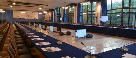 Une salle de conférence avec des rangées de chaises, une longue table couverte de documents et d’appareils électroniques, et de grandes fenêtres laissant entrer la lumière naturelle.