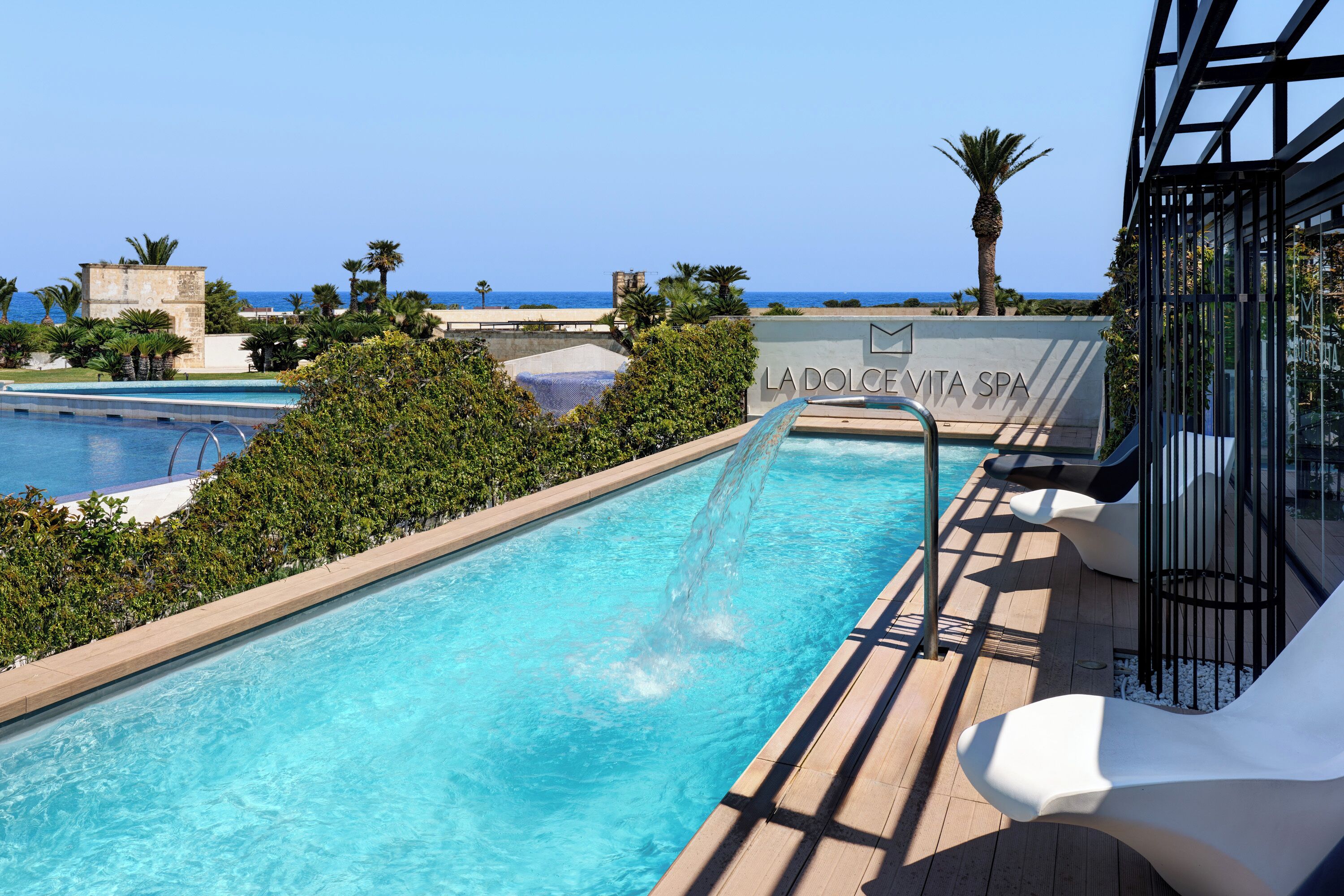 Ein Schwimmbecken mit Wasserrutsche, umgeben von Grün und Blick aufs Meer.