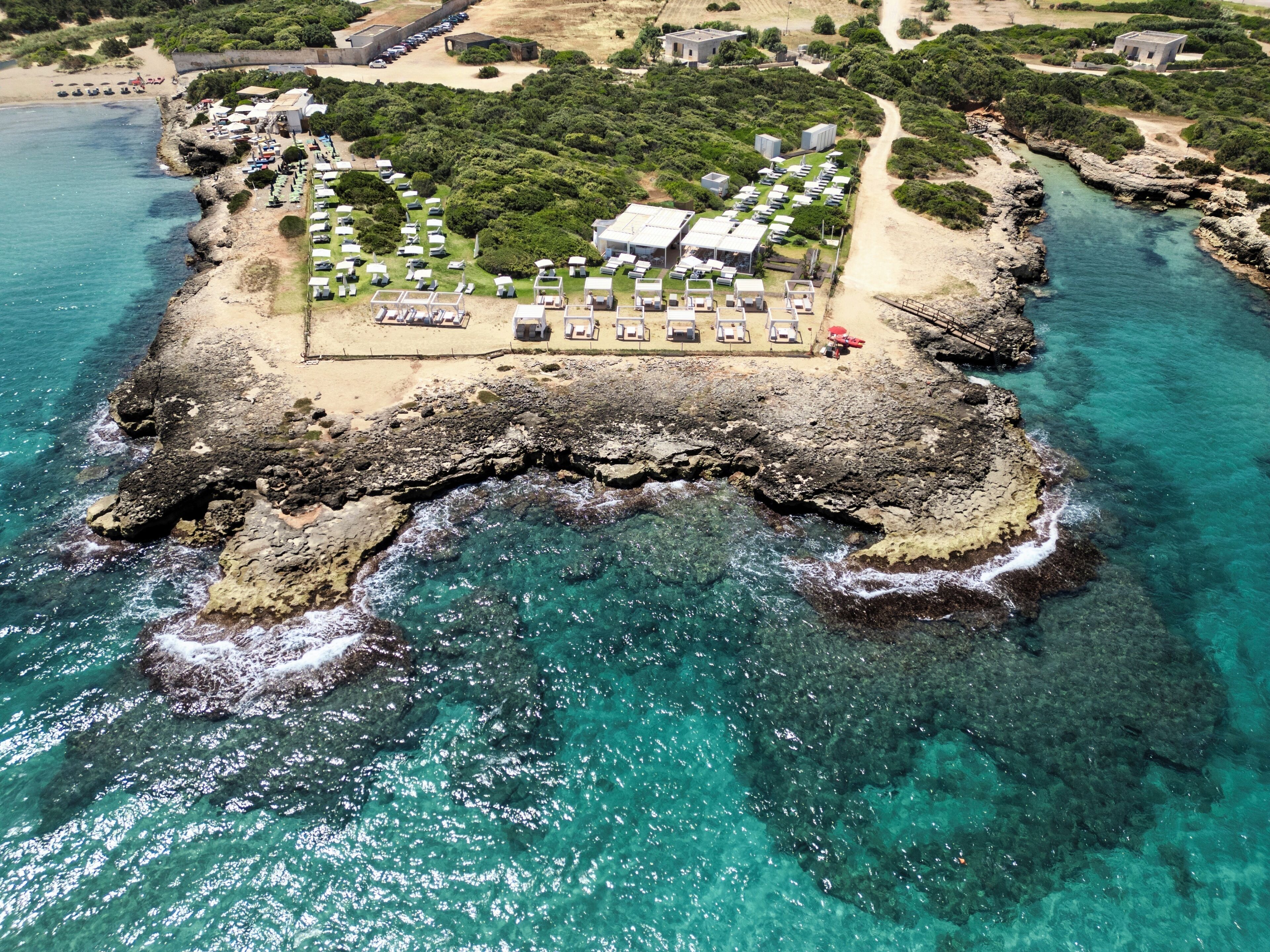An aerial view of a coastal property with a beach, rocky shoreline, and clear blue waters.