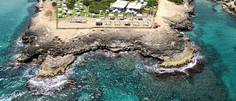 An aerial view of a coastal property with a beach, rocky shoreline, and clear blue waters.