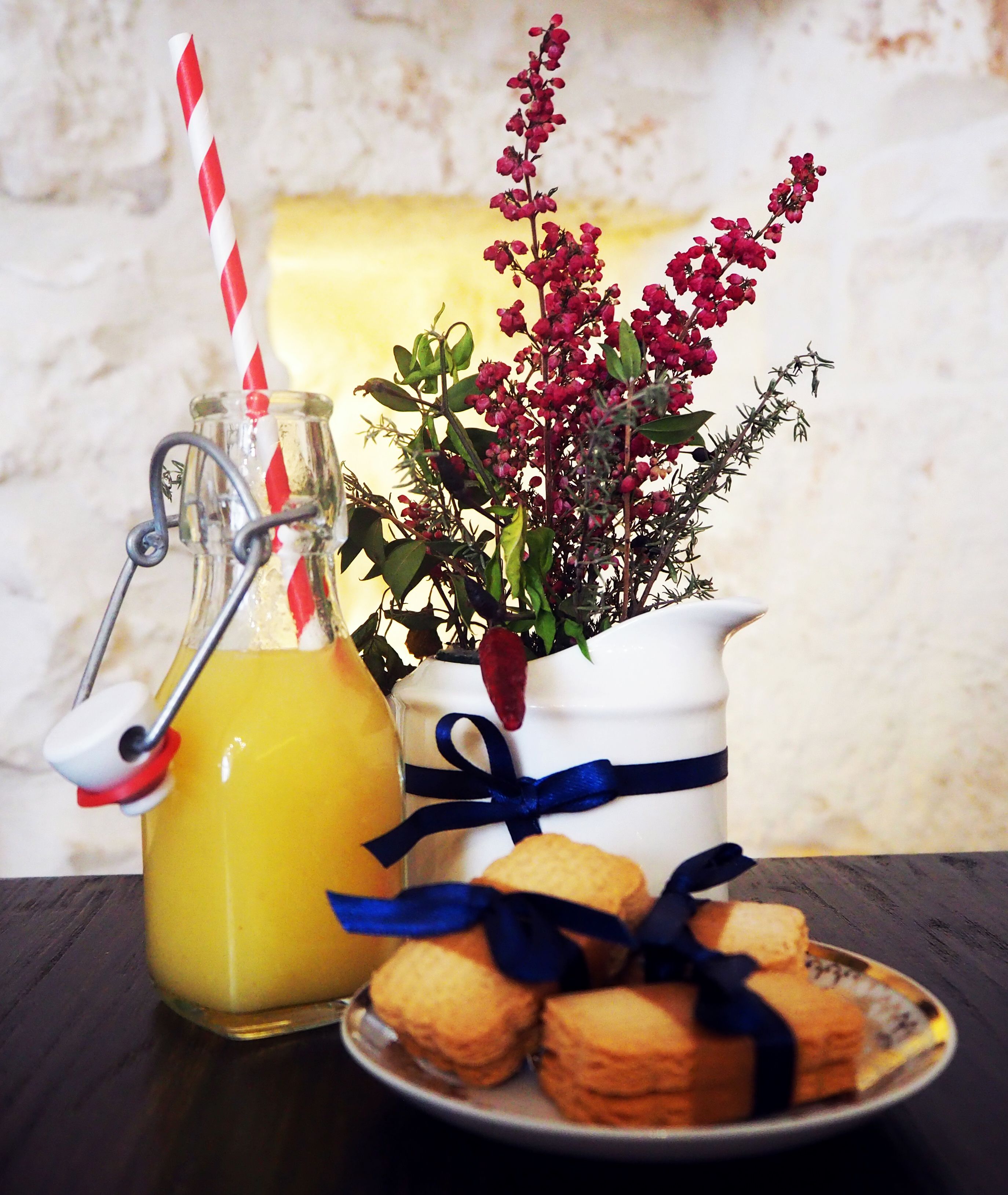 A glass bottle with a red and white striped straw, a white pitcher with a blue ribbon, and a plate of cookies.