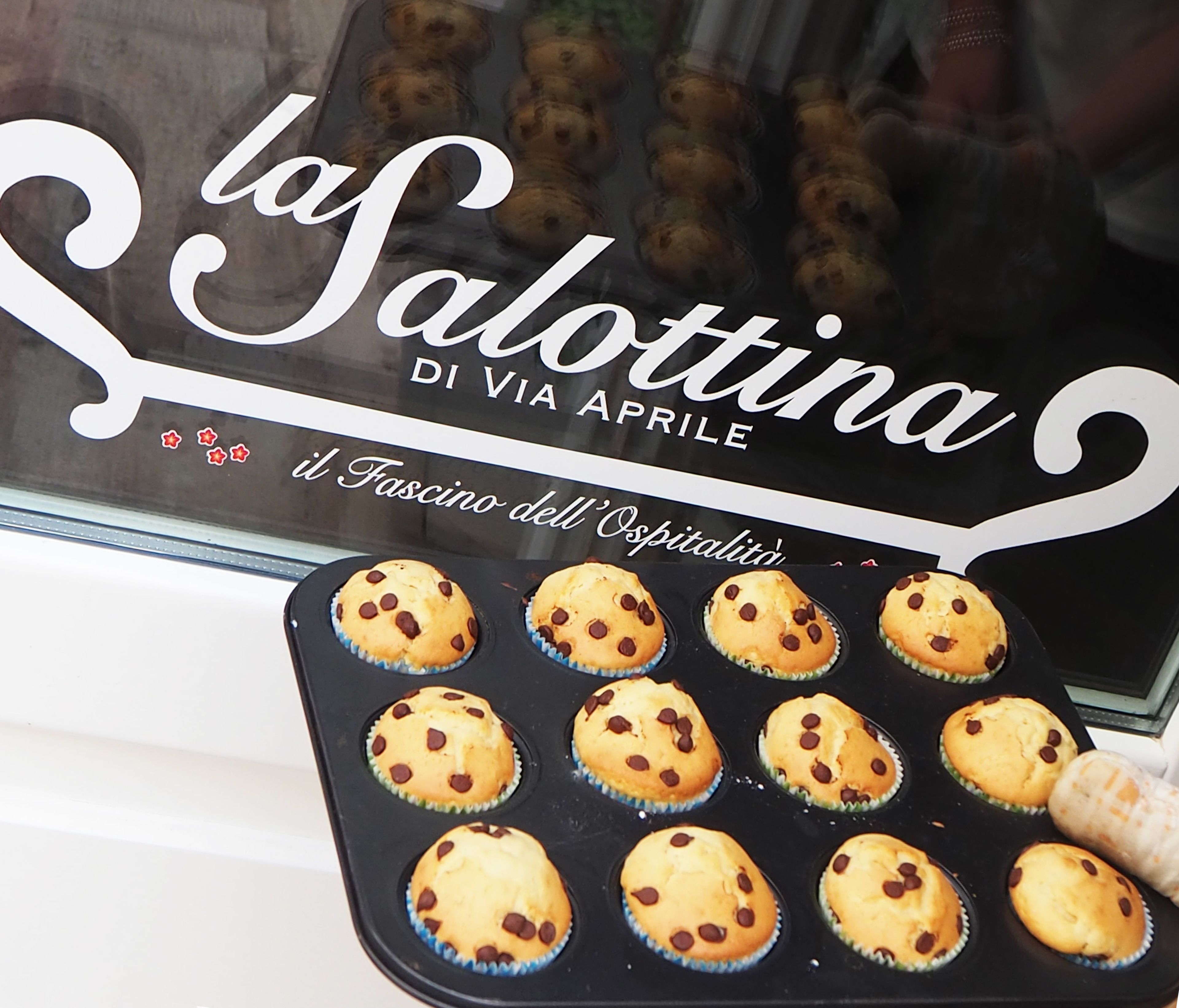 A bakery window with the sign "la Salottina DI VIA APRILE" and a display of freshly baked muffins.