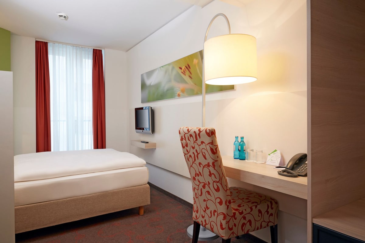 A hotel room with a bed, a desk with a telephone, a chair, a television, and a painting on the wall.