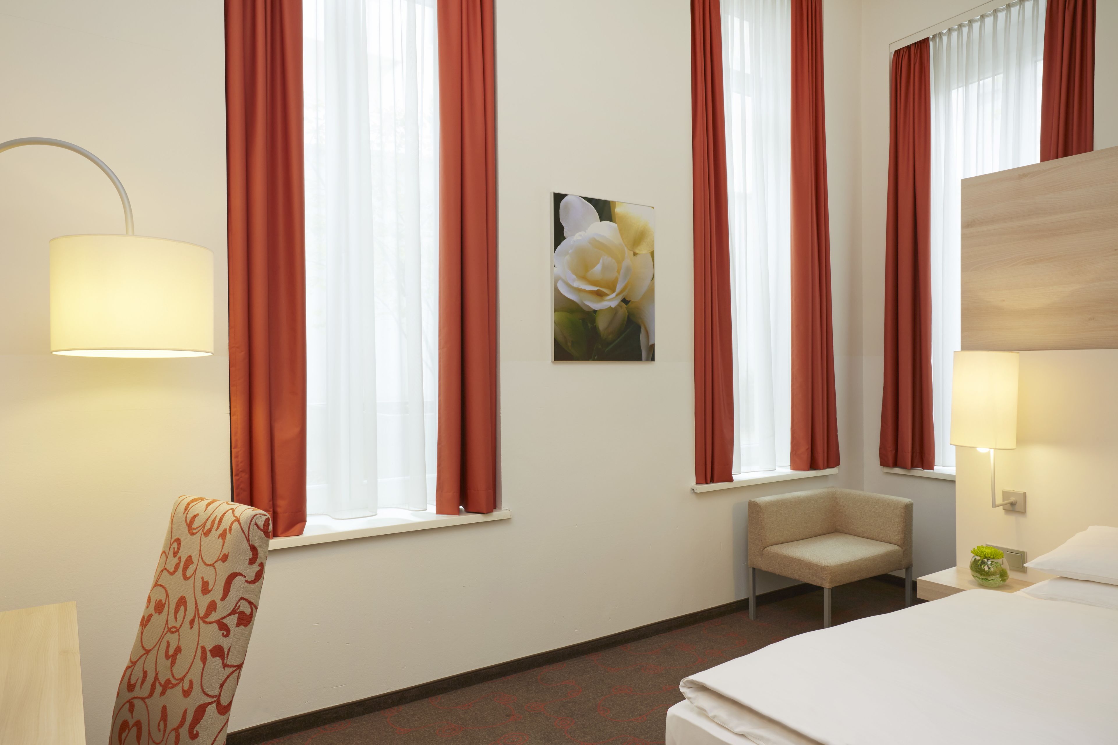 Une chambre d’hôtel avec un lit, une chaise, un bureau avec une lampe et un tableau représentant des fleurs.