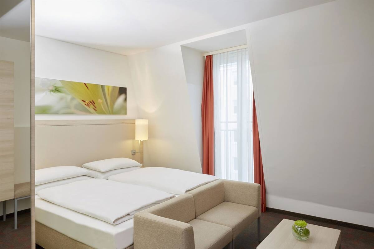 A hotel room with a bed, a sofa, a lamp, a window with curtains, and a painting on the wall.