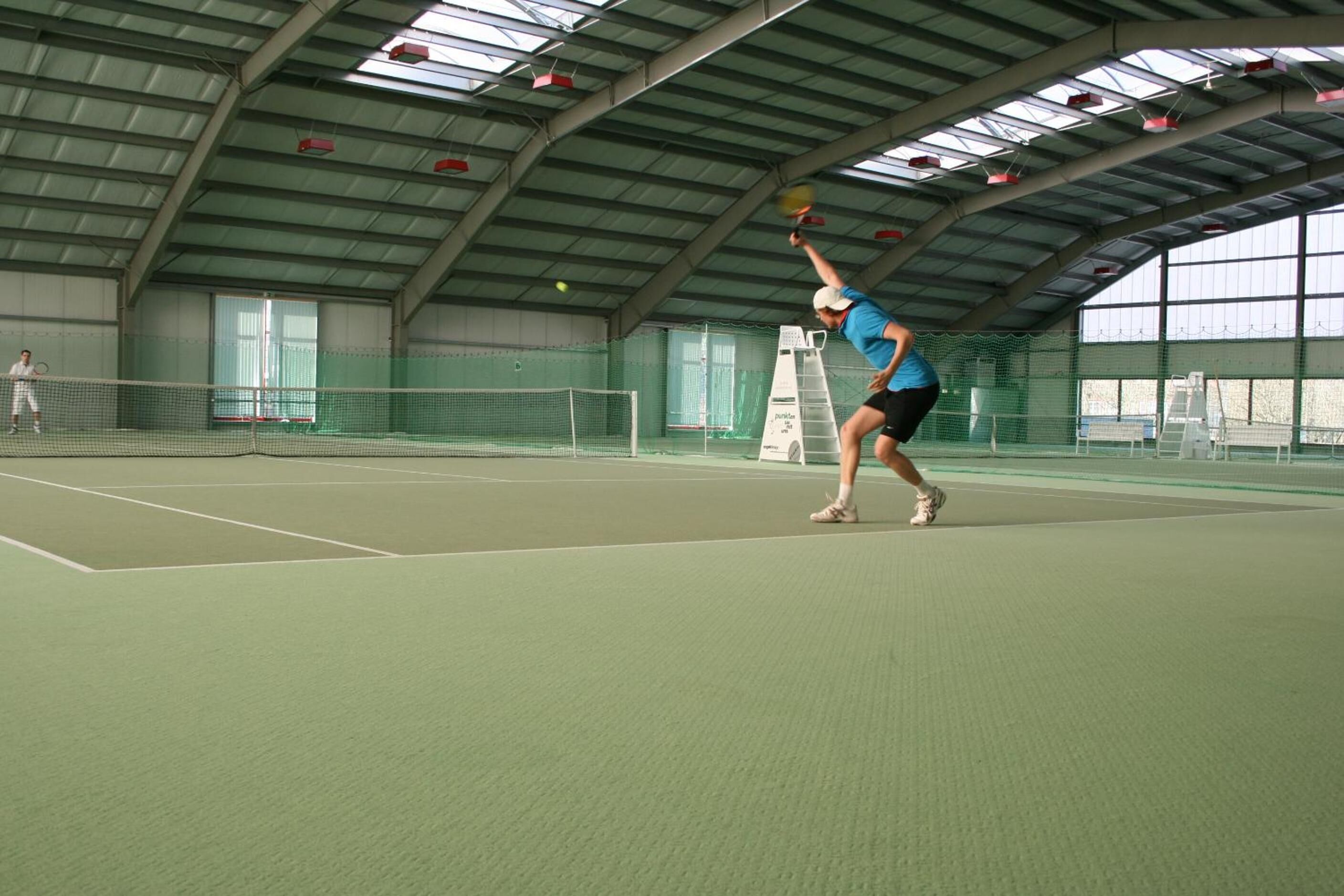 Una cancha de tenis interior con red, un jugador en movimiento y una escalera.