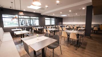 A modern dining area with multiple tables and chairs, large windows, and pendant lighting.