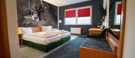 Una habitación de hotel con cama, escritorio, silla, una bicicleta colgada en la pared y ventana con persianas rojas.