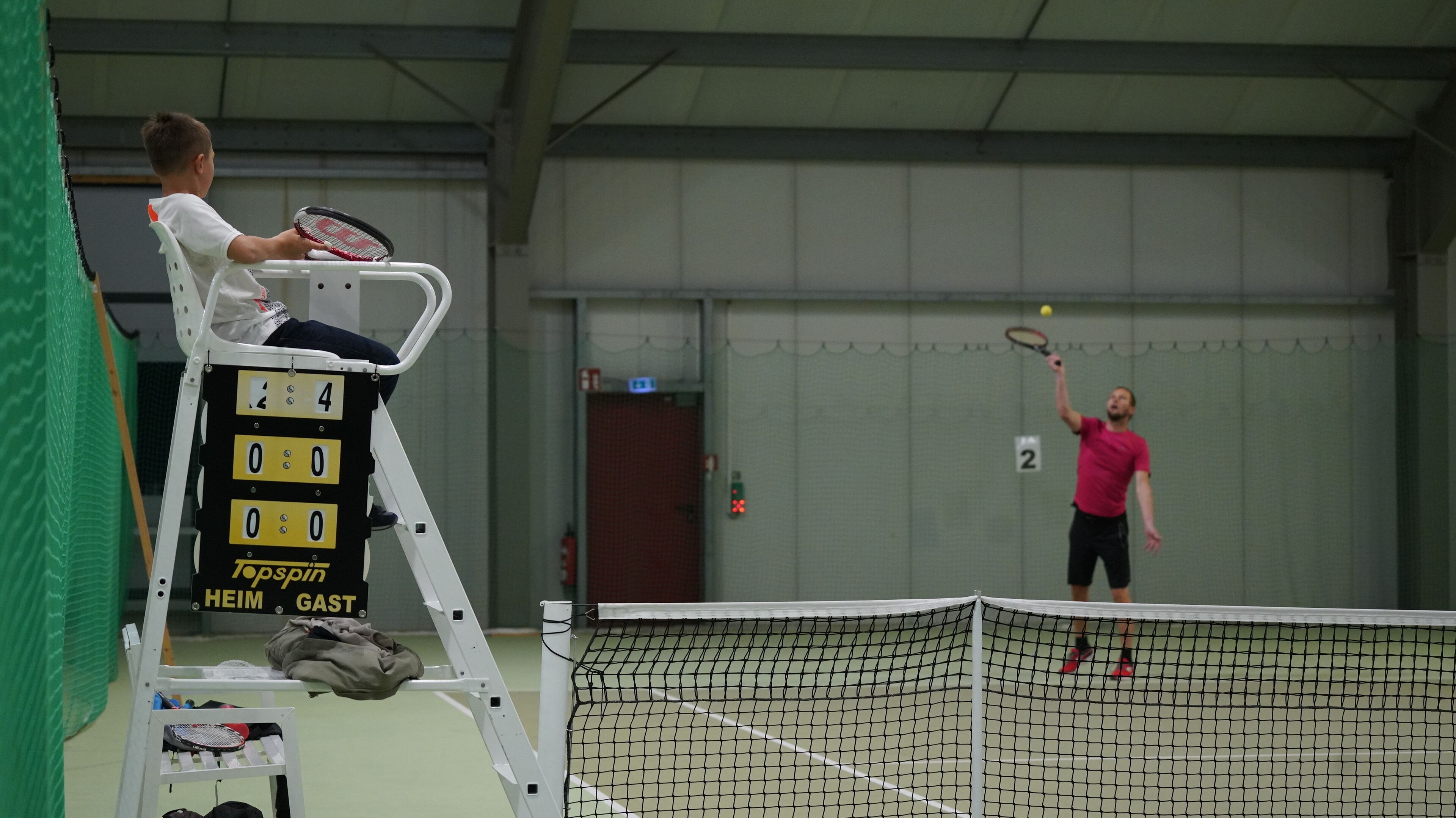 Un court de tennis avec un tableau d’affichage, un joueur qui effectue un service et un télésiège.