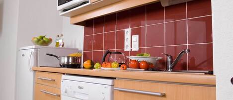 A compact kitchen with a microwave, stove, and sink. There are various fruits and vegetables on the counter, and a dish rack above the stove.